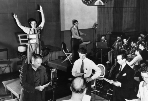Студия CBS во время знаменитой передачи. Орсон Уэллс за пультом, с поднятыми руками. На переднем плане остальные актёры, на заднем плане играет оркестр.